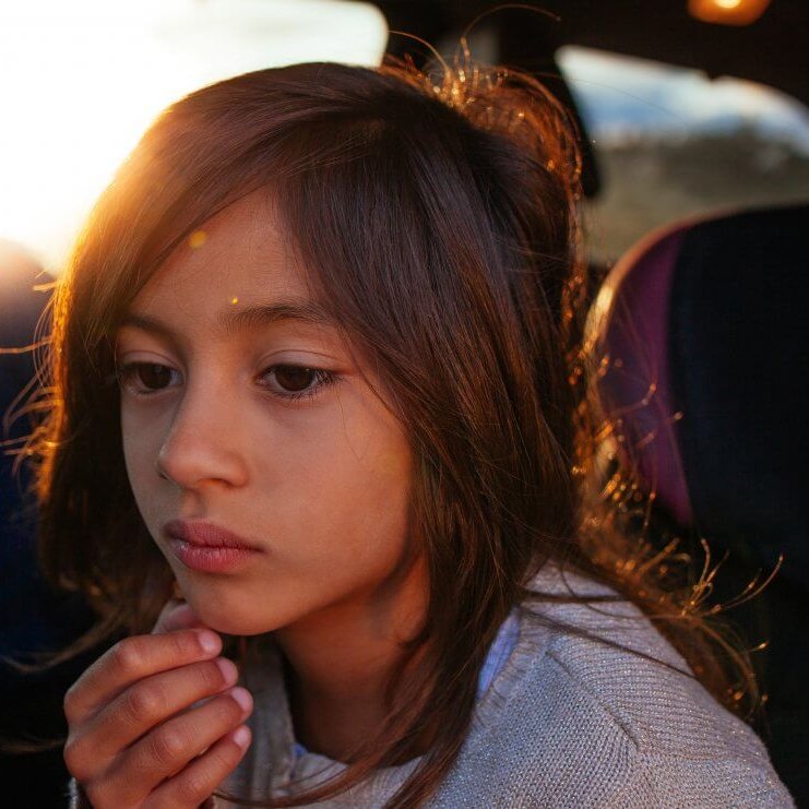 A child with long, brown hair and light brown skin sits in a car, close to the camera. The child is touching their chin with one hand and looking with an unfocused gaze into empty space. The sun is low in the sky and partially occluded by the child's head, giving them a golden highlight.